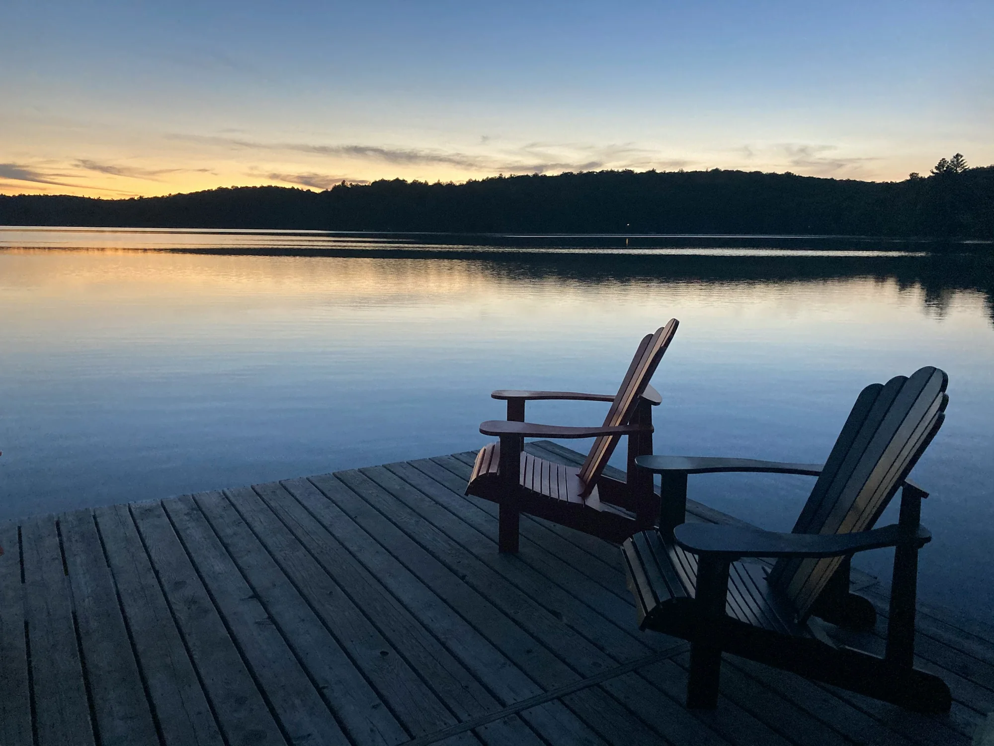 Two Muskoka chairs on a dock at sunset over a calm lake near Bracebridge
