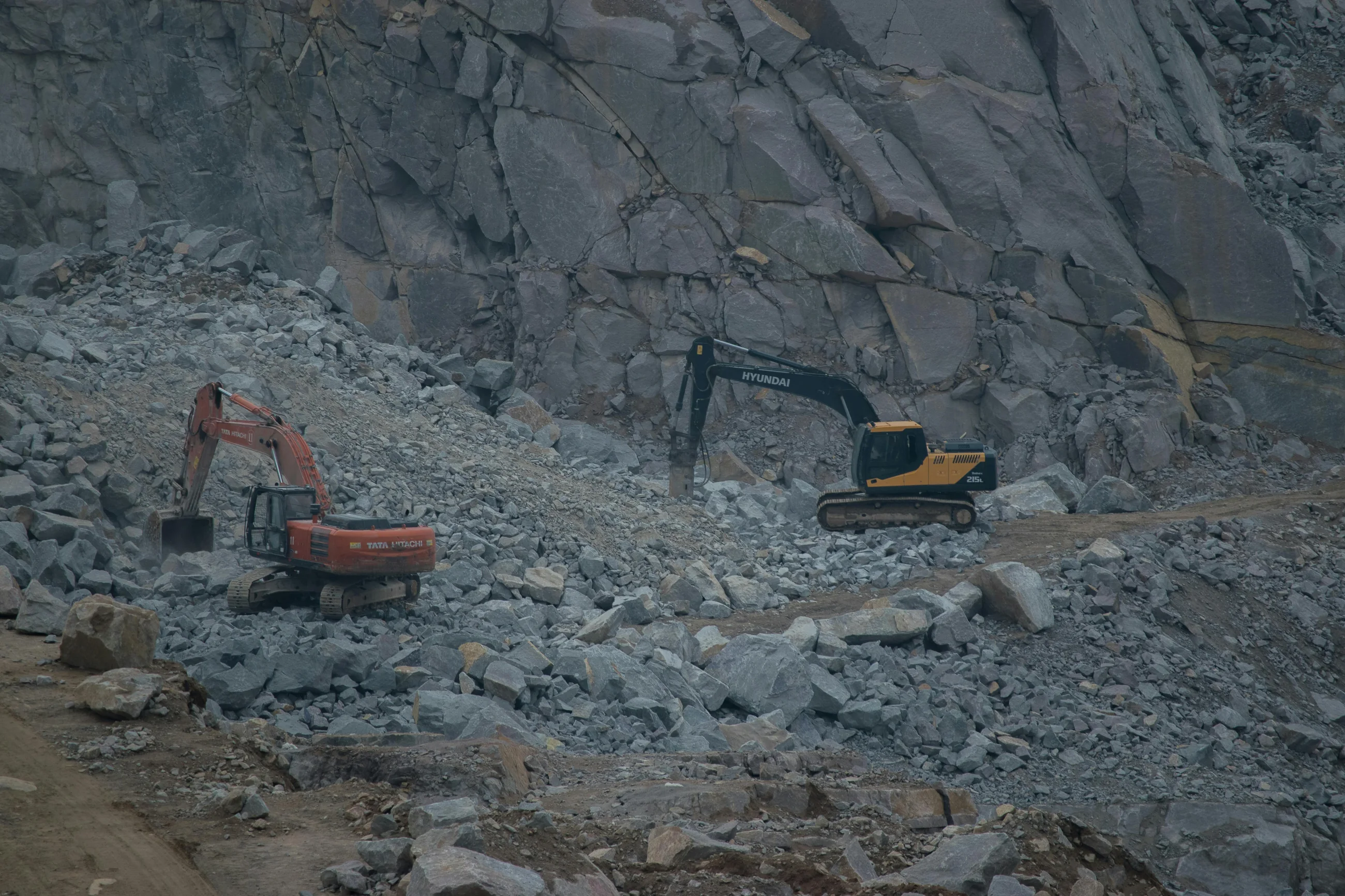 An excavator working a rock quarry site, dust rising against grey stone