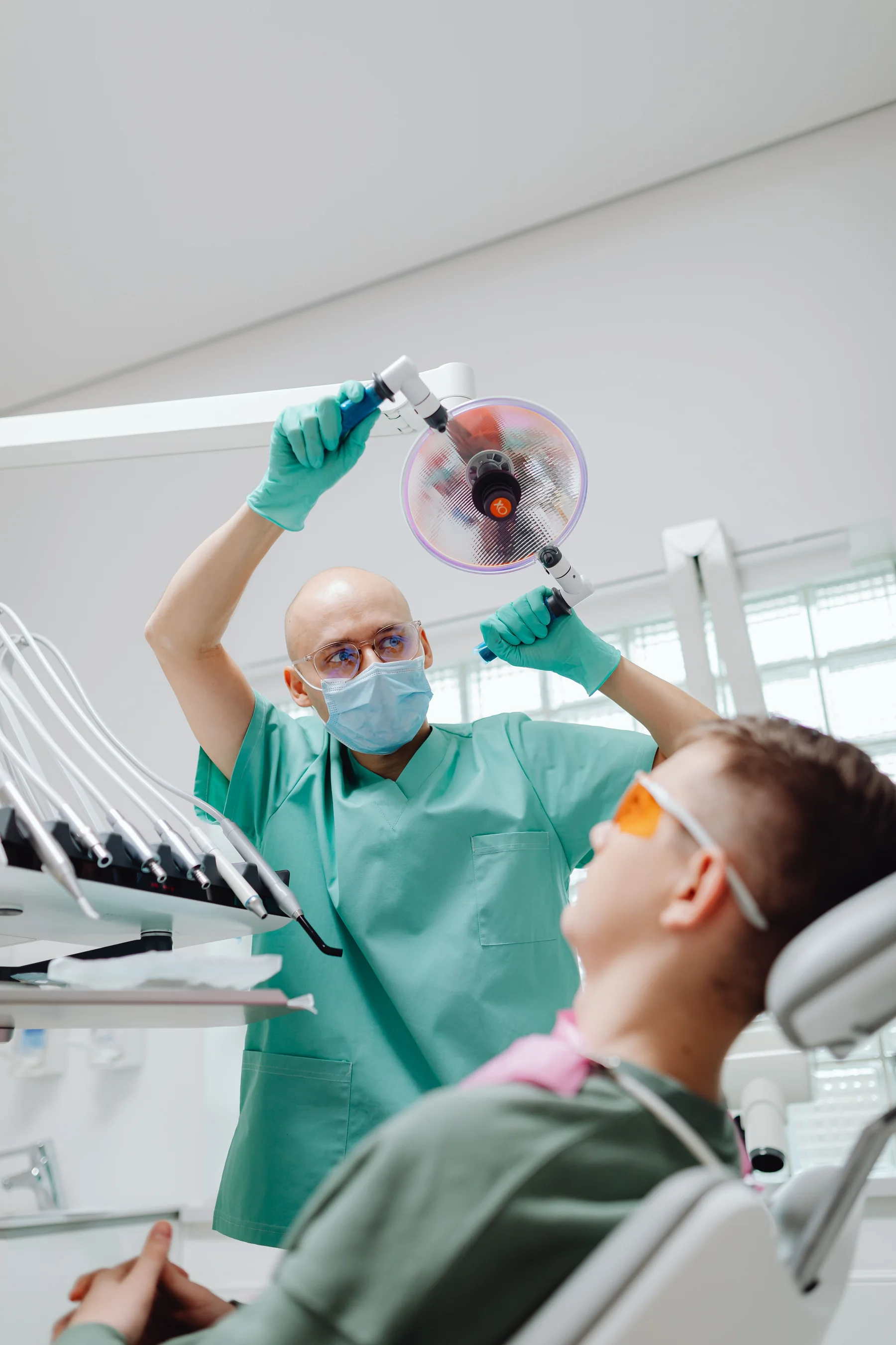 A dentist in green scrubs adjusting the overhead light above a patient in safety glasses