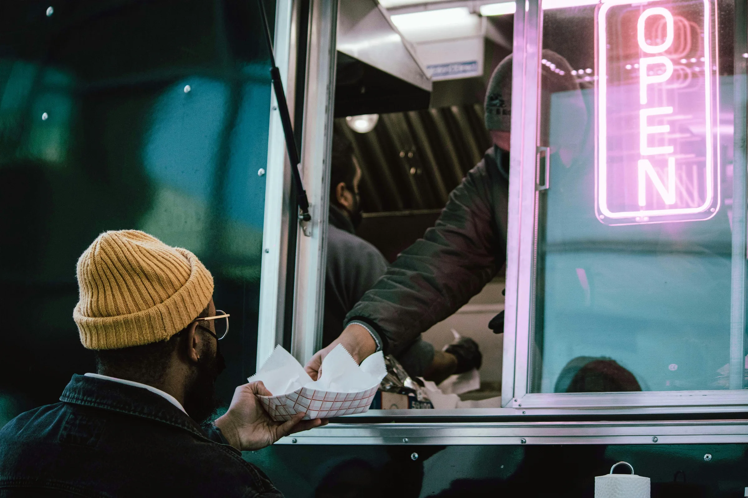 A customer receiving food through a food truck's glowing OPEN sign window at night