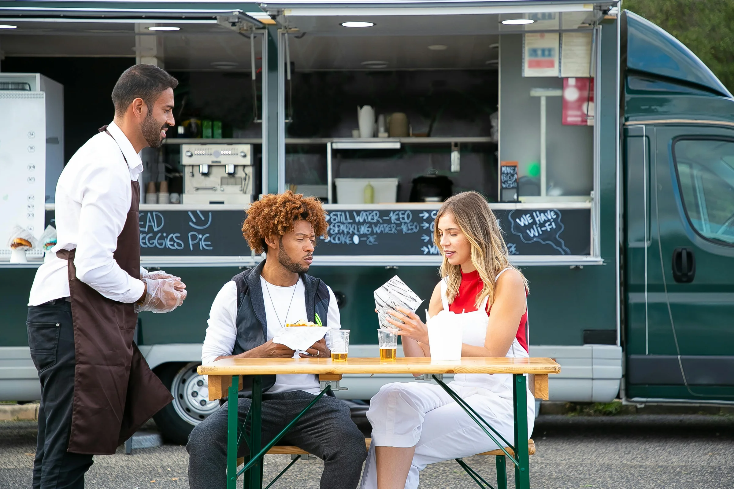 Customers seated at a picnic table being served by a food truck vendor on a sunny day