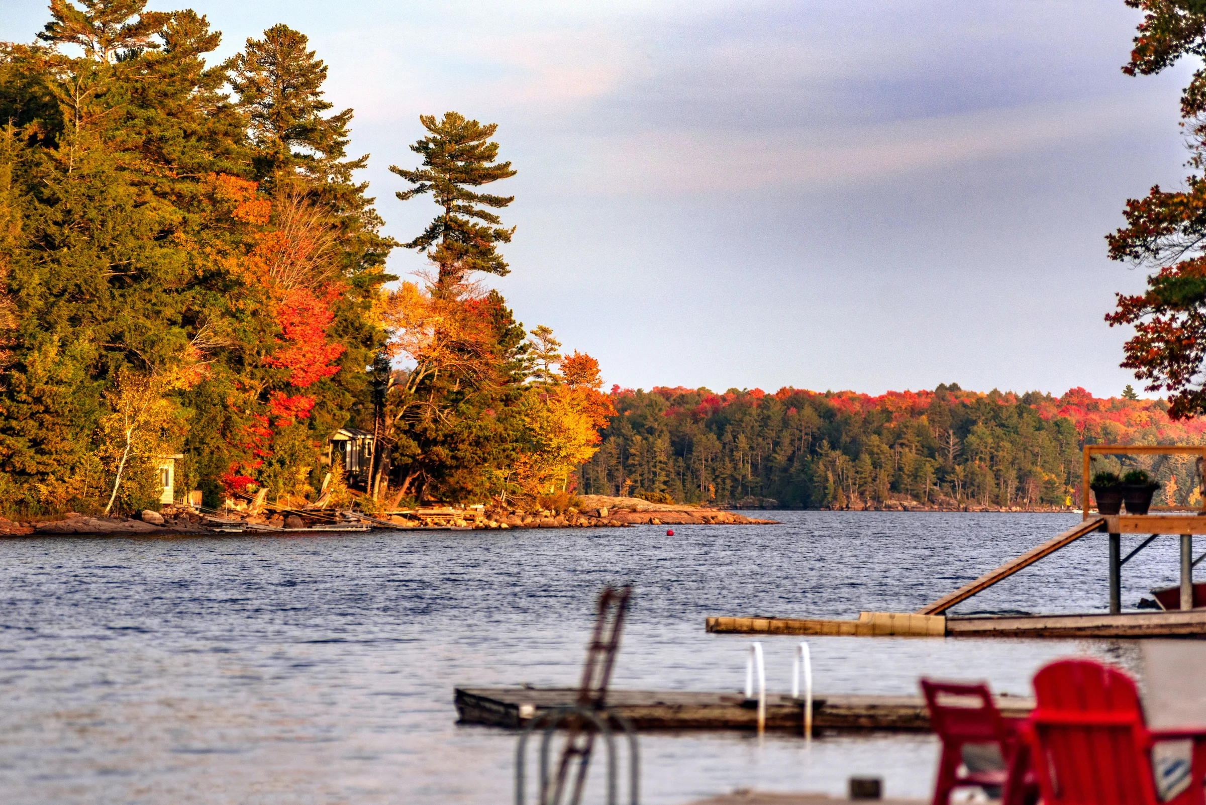 Red Muskoka chairs on a dock beside a lake in fall colour near Gravenhurst