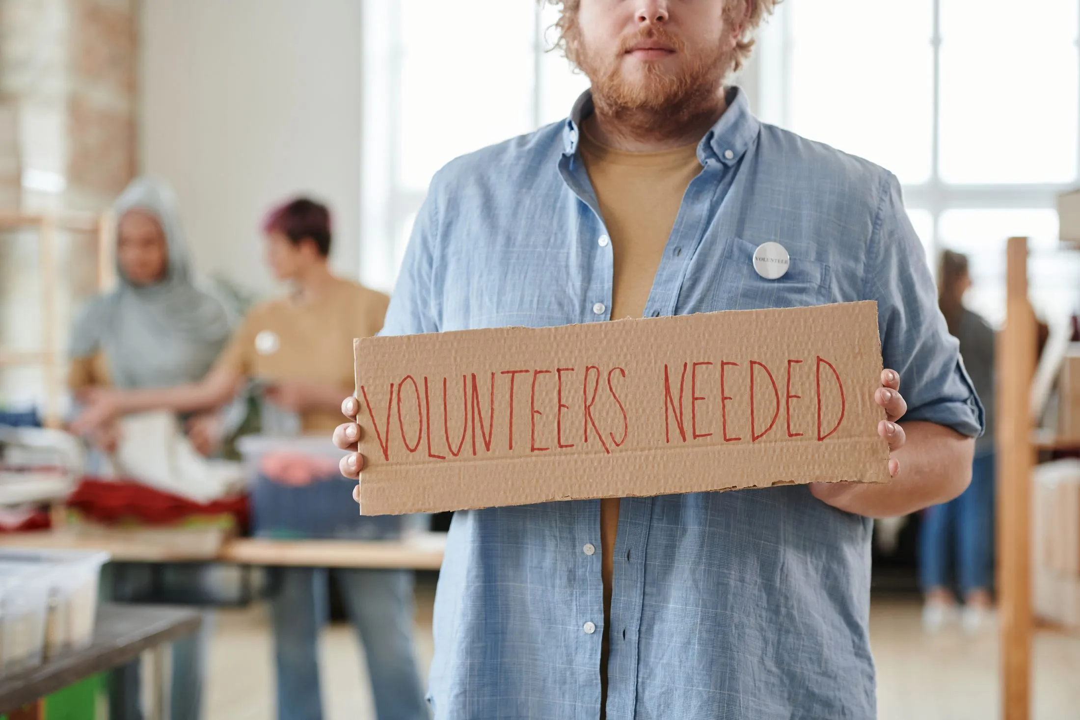 A man holding a 'Volunteers needed' sign in a community centre
