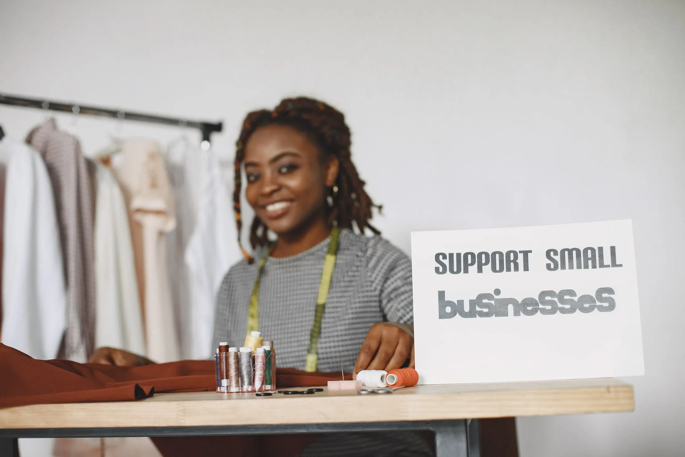 A seamstress smiling at her worktable beside a 'Support small businesses' sign