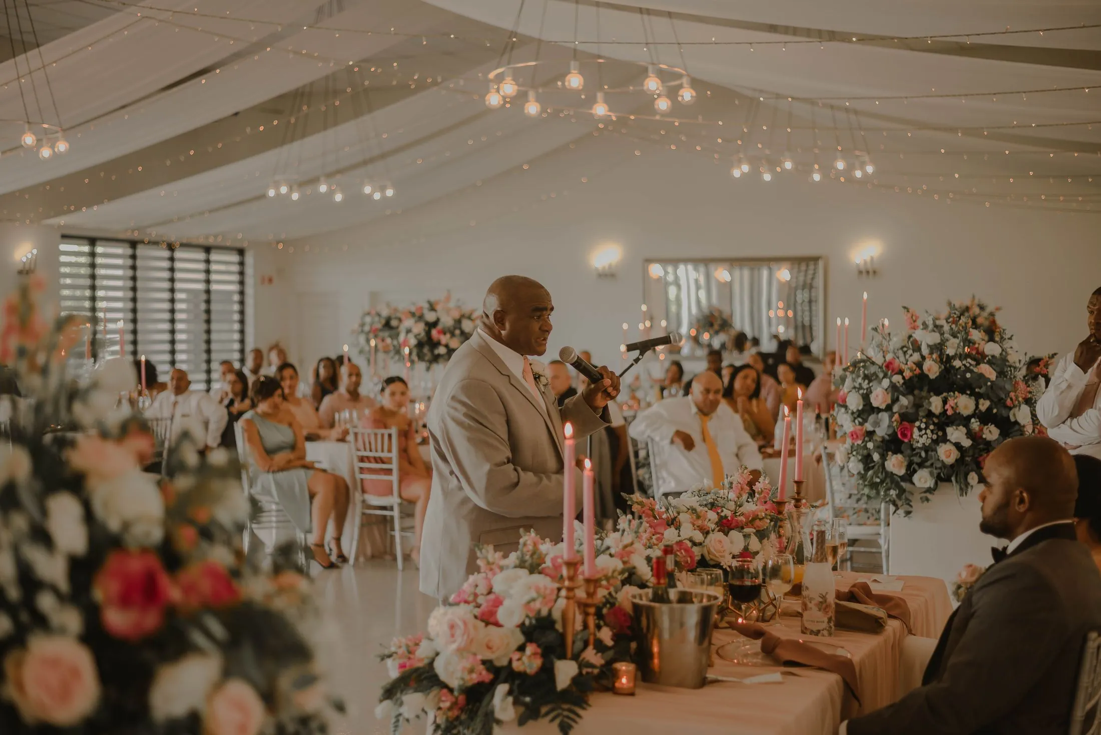 A wedding reception with floral centrepieces and guests listening to a speech